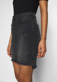 Černá deninová sukně s přiléhavým střihem, roztrhaným lemem a ošuntělými detaily. Obsahuje přední kapsu a rozparek na levé straně.