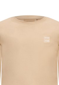 Beige Langarmshirt aus weichem Material; hat einen runden Ausschnitt und ein kleines, quadratisches weißes Logo-Patch auf der Brust.