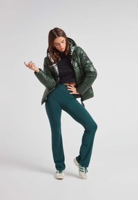 Chaqueta acolchada verde con capucha de logo en blanco y negro, combinada con pantalones azul claro y zapatillas blancas con rayas verdes; textura suave.