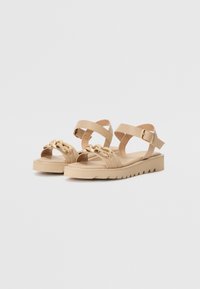 Beige sandaler med flate, teksturerte såler, justerbare ankelremmer og dekorativt hardware i gulltone som pryder fronten.