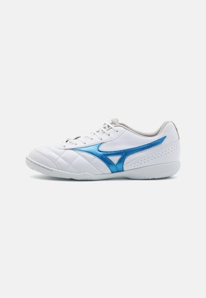 Zapatilla deportiva blanca con textura acolchada, acentos azules, parte superior de malla y suela de goma. Presenta un diseño de cordones y perforaciones para transpirabilidad.