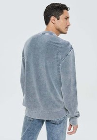 Pull en tricot gris avec une finition texturée, doté de poignets et d'un bas côtelés, et d'une coupe décontractée. Porté par-dessus un jean bleu.