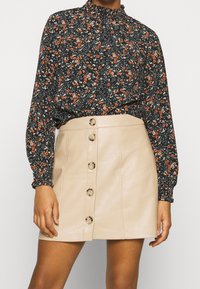 Blommig blus i svart med orange och vita blommor, hög hals och långa ärmar. Beig läder mini kjol med framknappar och slät textur.