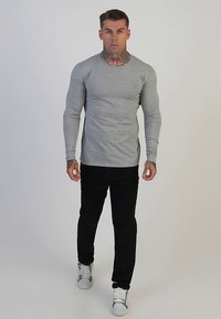 Grijze longsleeve shirt, aansluitend model, gladde textuur. Gecombineerd met zwarte broek en witte sneakers, staand op een effen achtergrond.