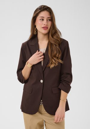 Femme aux longs cheveux ondulés portant un blazer marron foncé sur un haut clair, un pantalon beige, des boucles d'oreilles créoles en or et plusieurs bagues et bracelets en or.
