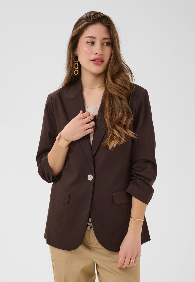 Femme aux longs cheveux ondulés portant un blazer marron foncé sur un haut clair, un pantalon beige, des boucles d'oreilles créoles en or et plusieurs bagues et bracelets en or.
