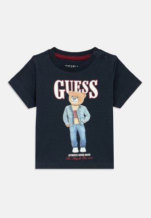 Maglietta blu navy per bambini piccoli con bottoni a pressione, con orso in piedi che indossa una giacca di jeans e pantaloni sotto la scritta grande "GUESS".
