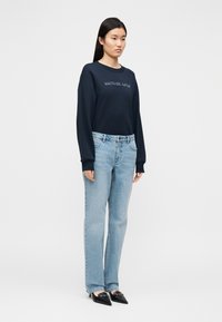 Tamsiai mėlynas džemperis su siuvinėtu logotipu, derinamas su šviesiai mėlynomis tiesiosiomis džinsų kelnėmis ir juodais aukštakulniais su smailiomis noselėmis, fone paprastas fonas.