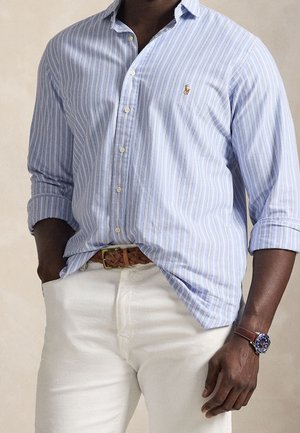 Homme portant une chemise boutonnée rayée bleu clair et blanc avec les manches retroussées, pantalon blanc, ceinture tressée marron et montre bleue avec bracelet marron.