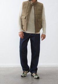 Uomo che indossa una camicia beige a maniche lunghe, un gilet imbottito marrone, jeans blu scuro e scarpe da trekking verdi, in piedi su un pavimento grigio contro un muro bianco.