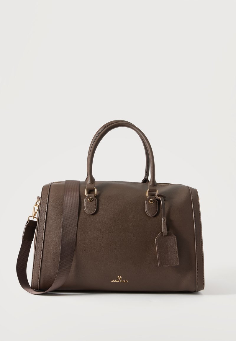 Sac de voyage en cuir brun foncé avec deux poignées, une bandoulière amovible et une étiquette à bagages sur un fond clair uni.