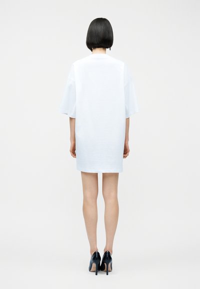 Γυναίκα με κοντά μαύρα μαλλιά, που φοράει ένα απλό, φαρδύ λευκό φόρεμα T-shirt και μαύρα ψηλοτάκουνα παπούτσια, στραμμένη πλάτη σε λευκό φόντο.