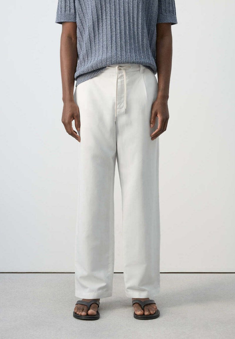 Personne portant un pantalon blanc ample avec taille à cordon, un haut en maille gris à manches courtes, et des sandales noires à boucle pour les orteils, debout sur un sol gris.