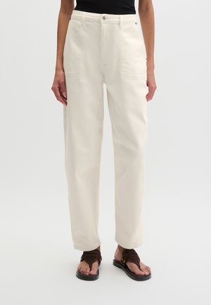 Personne portant un pantalon taille haute, coupe droite, couleur écrue, avec des poches plaquées et des sandales marron foncé sur un fond neutre.