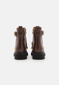 Botas de tobillo de cuero marrón con una suela negra texturizada, detalles en dorado y correas ajustables en la parte trasera. Detalle de tachuelas en los lados.