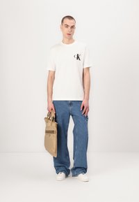 White short-sleeve t-shirt with black logo at chest, paired with loose blue denim jeans. The model holds a beige bag with two handles.