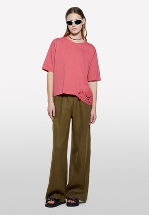 T-shirt corta rosa con dettagli in pizzo, abbinata a pantaloni larghi in lino verde oliva e sandali neri. Modella accessorizzata con occhiali da sole bianchi.