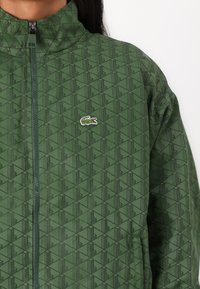Grün gemusterte Jacke mit hohem Kragen, die ein geometrisches Design aufweist und ein kleines, gesticktes Logo auf der Brust hat. Reißverschluss.