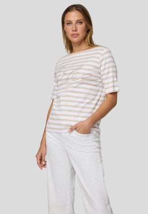 Frau trägt beige-weiß gestreiftes Kurzarmoberteil mit kursivem Text und weiße Hose, steht mit einer Hand in der Tasche vor grauem Hintergrund.