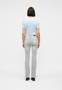 Γυναίκα που στέκεται γυρισμένη πλάτη, φορούσε εφαρμοστά τζιν ανοιχτού γκρι χρώματος, κοντομάνικο πουκάμισο με γαλάζιες και λευκές ρίγες και μαύρα παπούτσια με τακούνι.