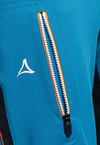 Kurtka z tkaniny w kolorze morskim z widocznym pomarańczowym zamkiem błyskawicznym, z wyraźnym białym logo i gładką fakturą. Zamek umieszczony z przodu.