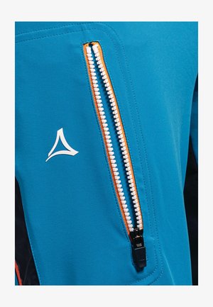 Jakke i blågrønt stof med en synlig orange lynlås, der har en fremtrædende hvid logo og en glat tekstur. Lynlåsen er placeret foran.