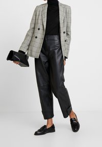 Doppelt geknöpfter Blazer in hellgrauem Hahnentritt, schwarzer Rollkragenpullover, Lederhose, schwarze Clutch und Slipper mit Hardware-Akzenten.