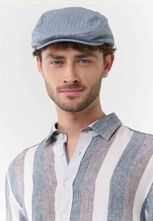 Man with short curly brown hair and beard wearing light blue newsboy cap and vertical striped button-up shirt in soft colors.