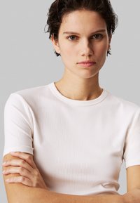 Weißes, geripptes T-Shirt mit kurzen Ärmeln und rundem Ausschnitt. Der Stoff wirkt weich und leicht strukturiert. Das Model hat die Arme vor der Brust verschränkt.