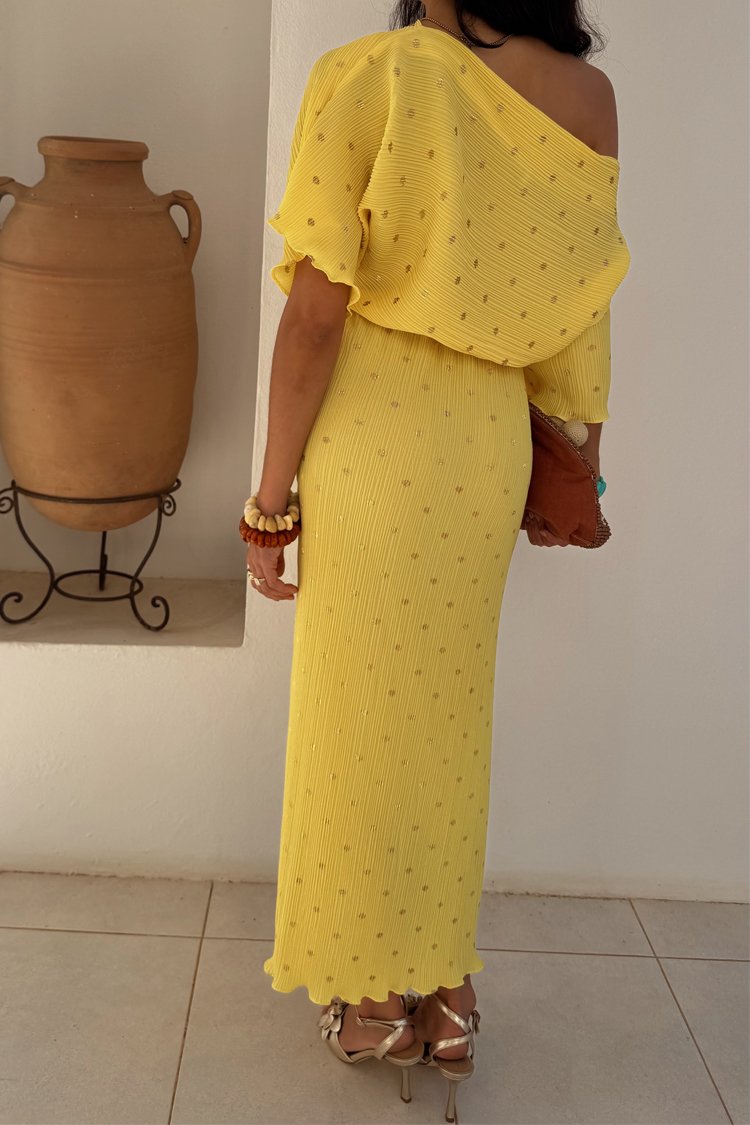 Żółta sukienka z prążkowanym wzorem i odkrytymi ramionami, ozdobiona małymi złotymi akcentami i falbaniastym dołem. Połączona z metalicznymi szpilkami.