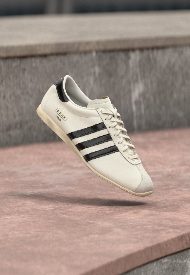 Λευκό αθλητικό παπούτσι Adidas με μαύρες ρίγες και μπεζ σόλα, αιωρούμενο πάνω από ροζ σκυρόδεμα και γκρι πέτρινο φόντο.