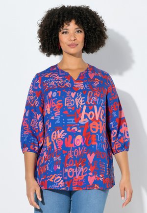 Kvinde med krøllet hår iført en blå bluse dækket af forskellige røde og lyserøde "kærlighed"-tekster og hjerte-mønstre, kombineret med lyseblå jeans.