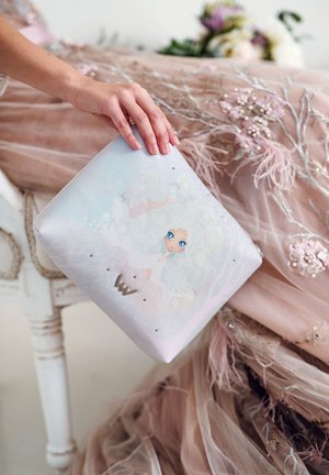 Pastel-colored pouch featuring a whimsical illustration, silver accents, and embellishments. Hand positioned on the pouch, set against a soft fabric background.