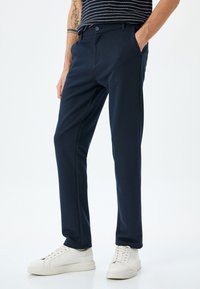 Marineblauwe broek van een gladde stof met een slim fit, voorzien van zijzakken en een knoopsluiting, gecombineerd met witte sneakers.