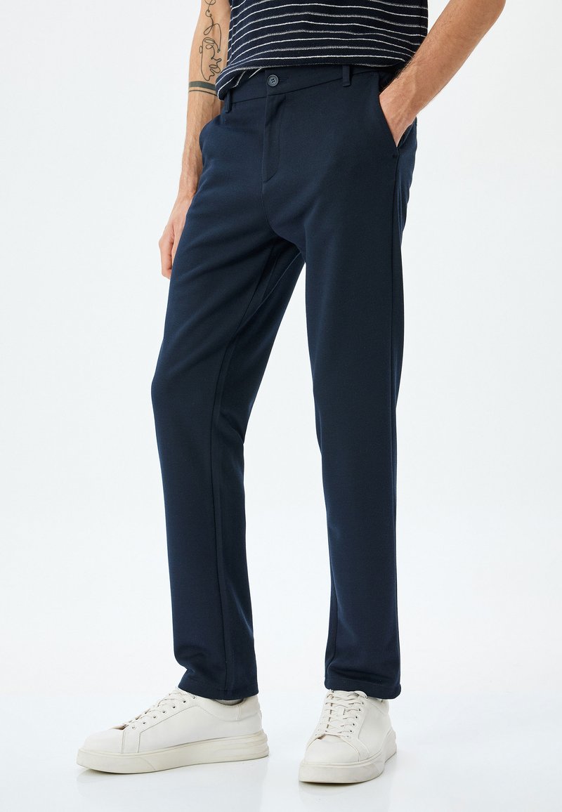 Marineblauwe broek van een gladde stof met een slim fit, voorzien van zijzakken en een knoopsluiting, gecombineerd met witte sneakers.