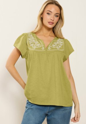 Kobieta ubrana w jasnobeżową bluzkę z krótkim rękawem ozdobioną białą kwiatową haftowaną częścią na piersi, w połączeniu z niebieskimi dżinsami, pozująca na tle beżowej ściany.