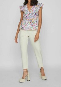 Femme portant un pantalon blanc taille haute, des talons compensés crème et un chemisier à manches courtes floral avec des épaules à volants.