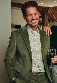 Olijfgroene corduroy blazer over een lichtgroen bloemenoverhemd met knoopdetails; houdt een glas rode wijn vast.
