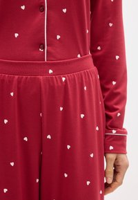 Ensemble de pyjama rouge à manches longues avec motifs de cœurs blancs, passepoil blanc sur les poignets et la zone des boutons, et pantalon à taille élastique.