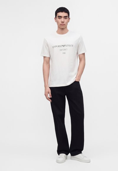 Νέος άνδρας με λευκό t-shirt με το λογότυπο Emporio Armani, μαύρο παντελόνι και λευκά αθλητικά παπούτσια στέκεται μπροστά σε απλό φωτεινό φόντο.