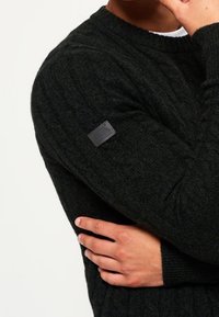 Maglione a coste verde scuro con scollo rotondo, dotato di un piccolo etichetta nera sulla manica sinistra e polsini a costine.