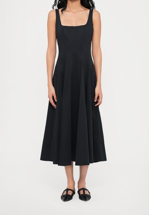 Kobieta w czarnej, bezrękawowej sukience midi z kwadratowym dekoltem i czarnych butach w szpic stoi na tle białego tła.