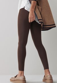 Braune Leggings aus elastischem Stoff, figurbetontes Design, glatte Textur, kombiniert mit einem beigen Slipper und geschichtet mit einem grauen Oberteil sowie einer braunen Strickjacke.