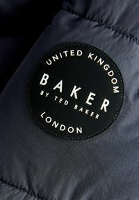 Etiqueta de tela circular negra con texto en relieve: "BAKER by TED BAKER", rodeada por "REINO UNIDO" y "LONDRES", sobre un material texturizado oscuro.