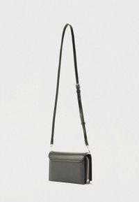 Borsa a tracolla in pelle nera con tracolla regolabile e tasca laterale con zip, appesa su uno sfondo grigio chiaro.