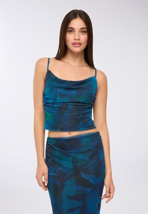 Giovane donna con lunghi capelli lisci che indossa un top senza maniche con motivo astratto blu e verde e una gonna coordinata, su sfondo bianco.