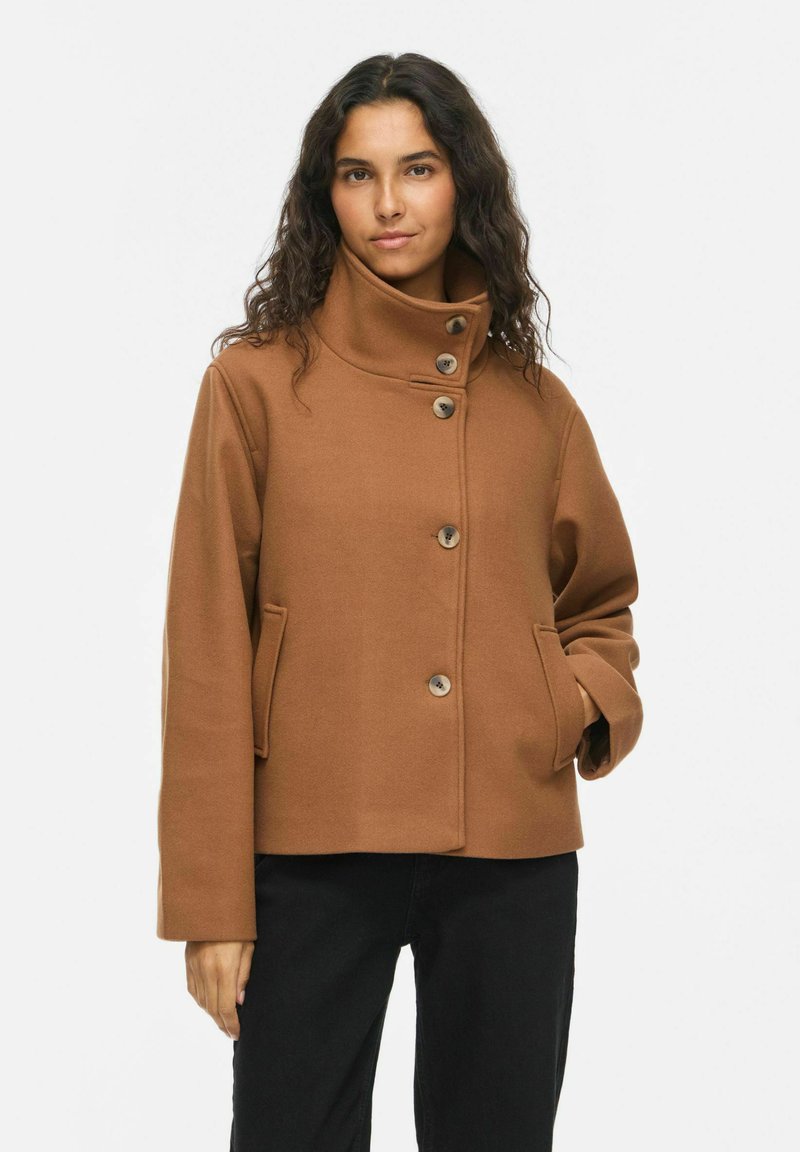 Femme aux longs cheveux bouclés et foncés, portant un manteau marron boutonné à col montant et un pantalon noir, debout devant un fond clair uni.