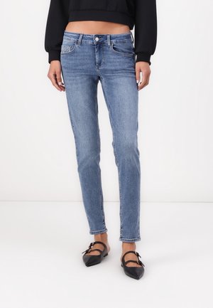 Kobieta ubrana w dopasowane jasnoniebieskie dżinsy, czarny krótki sweter oraz czarne spiczaste baleriny z podwójnymi paskami i klamrami.