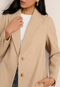 Beige wollen mix jas met een getailleerde pasvorm, notched revers, twee knopen en zijzakken. Draag over een witte geribbelde top. Subtiele textuur.