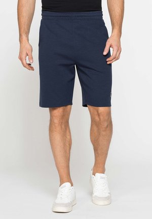 Uomo con pantaloncini blu navy e scarpe da ginnastica bianche in piedi davanti a uno sfondo chiaro e semplice.